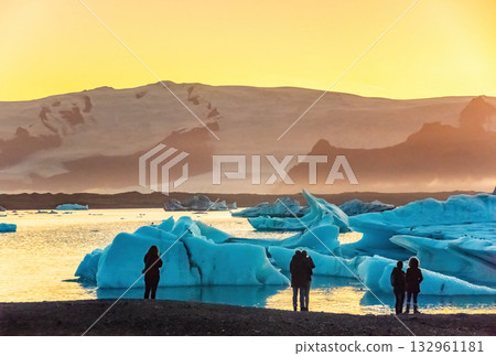 Tourists admire the stunning sunset over Iceland's Glacier Lagoon, surrounded by large icebergs. The serene scene captures the beauty of nature 132961181