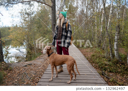 Glad woman and Vizsla dog enjoy quiet walk on ecological trail by lake in autumn nature park 132962497