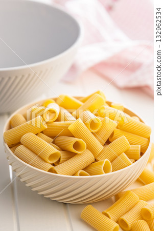 Uncooked rigatoni pasta in bowl on white table. Uncooked rigatoni pasta in bowl on white table. 132962534