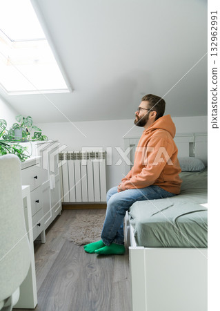 Man sitting alone on bed near window in bright room. Visual concept of morning routine, reflection, calmness and solitude in domestic environment. 132962991