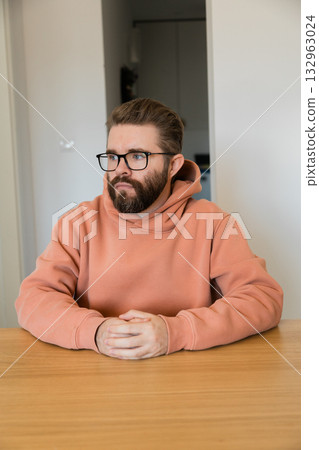 Man sitting at wooden table in apartment interior. Professional portrait of thoughtful male person symbolizing concentration, patience and waiting during communication or decision process. Man sitting at wooden table in apartment interior. Professional portrait of thoughtful male person symbolizing concentration, patience and waiting during communication or decision process. 132963024