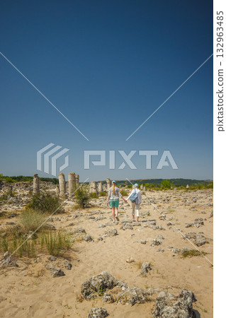Two women walking hand in hand among the ancient limestone columns of Pobiti Kamani, the Stone Desert near Varna, Bulgaria, under a clear summer sky. 132963485
