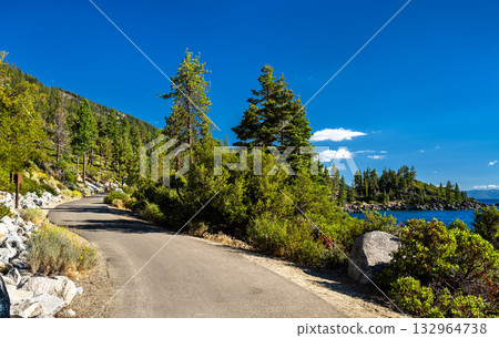 The paved Lake Tahoe East Shore Trail on a sunny day. The path curves past green trees and bushes, with the blue lake visible on the right. Represents outdoor recreation, hiking, and biking 132964738