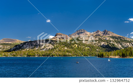 A scenic view of the deep blue Caples Lake in California, with the Sierra Nevada mountains rising in the background under a clear sky. A small boat and sailboat are visible on the water 132964752