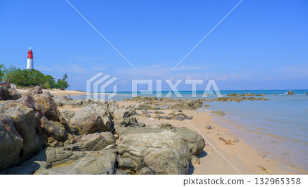 Red and White Lighthouse on a Tropical Shoreline with Sandy Beach and Rough Foreground Rocks 132965358