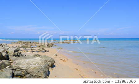 Wide Tropical Beach View with Sandy Shore, Foreground Rocks, and Calm Blue Ocean Under Bright Sky 132965360