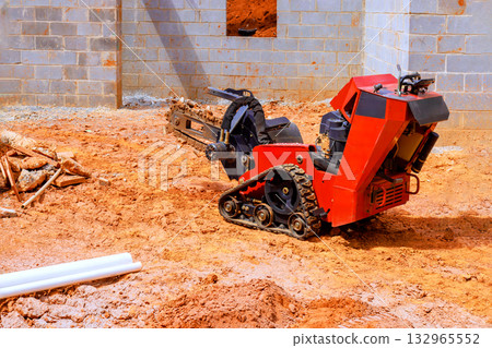 Compact trenching machine digs into earth at construction site, surrounded by unfinished walls under work day Compact trenching machine digs into earth at construction site, surrounded by unfinished walls under work day 132965552