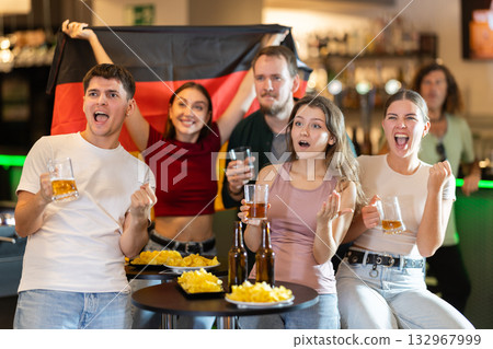 Group of fans of Germany team with a flag are watching the match on TV in beer bar. They are worried and supporting their Group of fans of Germany team with a flag are watching the match on TV in beer bar. They are worried and supporting their 132967999
