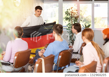 Young man tells his classmates the history of the state of Germany, holding national flag of Germany in his hands Young man tells his classmates the history of the state of Germany, holding national flag of Germany in his hands 132968205