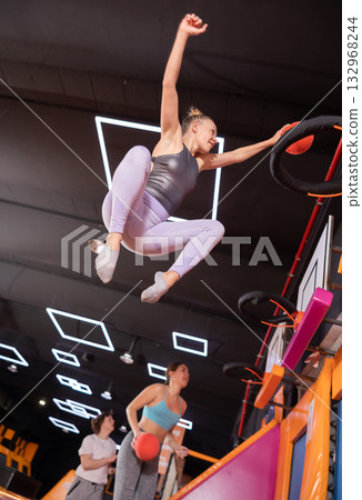 Active European female in sportswear playing basketball while bouncing on colorful trampolines indoors in leisure sports center 132968244
