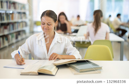 Female student reading book and taking notes in library Female student reading book and taking notes in library 132968336