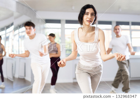 Happy girl and group of young active sport people practicing vogue dancing movements while training twist or charleston in dance hall Happy girl and group of young active sport people practicing vogue dancing movements while training twist or charleston in dance hall 132968347