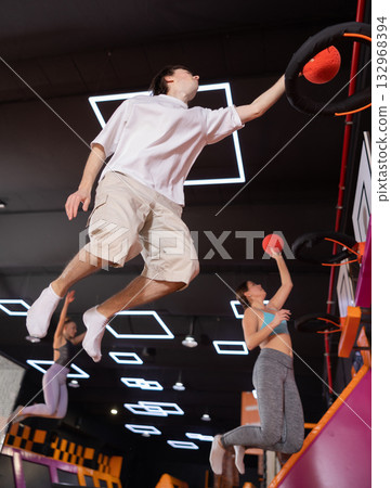 Active European young male in sportswear playing basketball while bouncing on colorful trampolines indoors in leisure sports center 132968394