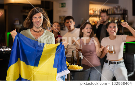 Company of sports fans waving flag of Sweden and supporting national team with beer in bar Company of sports fans waving flag of Sweden and supporting national team with beer in bar 132968404