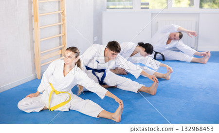 Young woman wearing kimono practicing leg stretching position in sports hall Young woman wearing kimono practicing leg stretching position in sports hall 132968453