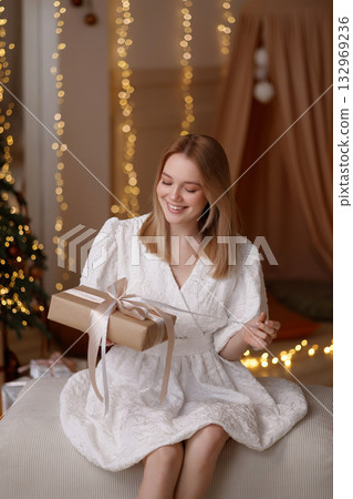 Young woman in white dress holding Christmas gift box indoors with fairy lights and beige curtain 132969236
