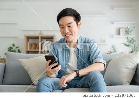 Smiling handsome young Asian man sitting on cozy couch Smiling handsome young Asian man sitting on cozy couch 132971365