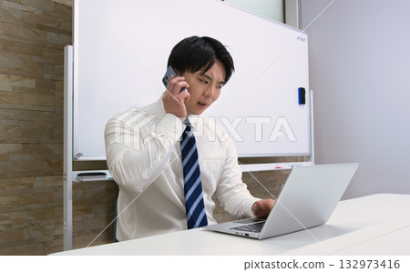 一位年輕的商人在辦公室裡一邊看著電腦一邊用智慧型手機聊天 132973416