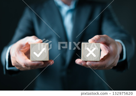 A businessman in a suit presents two wooden blocks, one with a white checkmark and the other with a white X, symbolizing decision making, evaluation, approval, rejection, and business choices. 132975620