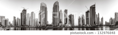 Monochrome city skyline with tall skyscrapers reflecting on calm water under a cloudy sky, Dubai Marina bay UAE 132976848