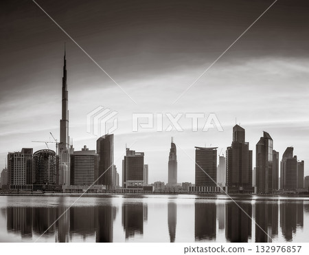 Monochrome city skyline with tall skyscrapers reflecting on calm water under a cloudy sky, Dubai Marina bay UAE 132976857