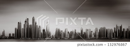 Monochrome city skyline with tall skyscrapers reflecting on calm water under a cloudy sky, Dubai Marina bay UAE 132976867