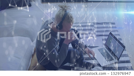 Working man wearing sweater typing on laptop in bedroom on rug, with mug headset digital overlay 132976947