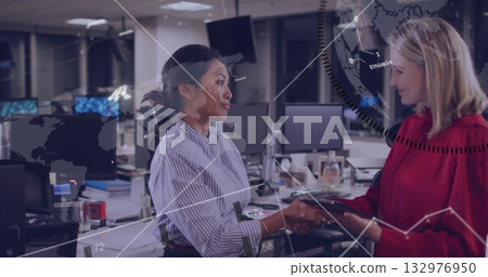 Businesswoman in red blouse handing tablet to colleague in striped shirt in office, with monitors 132976950