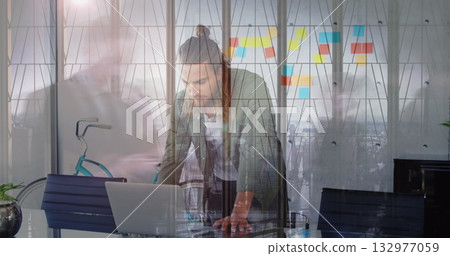 Leaning bearded man in beanie working on laptop at glass office table, with sticky notes, bicycle 132977059