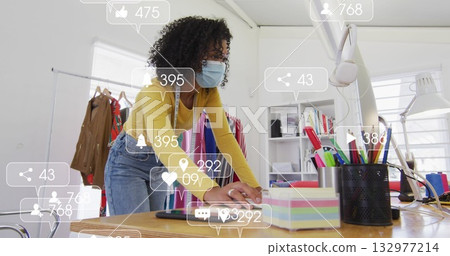 Leaning designer wearing yellow top face mask using tape measure at studio desk, with clothing rack Leaning designer wearing yellow top face mask using tape measure at studio desk, with clothing rack 132977214