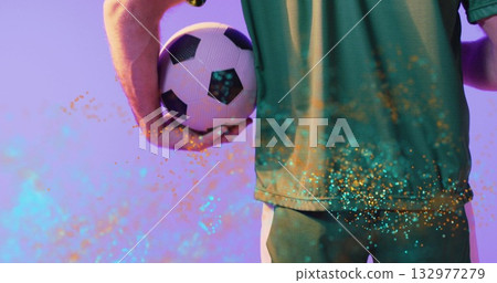Athlete wearing green jersey and shorts holding soccer ball behind waist in studio, with confetti 132977279