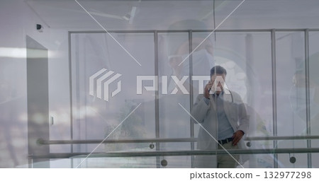 Using smartphone senior doctor in white coat speaking through glass partition at hospital corridor 132977298