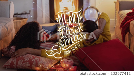 Cuddling couple wearing sweatshirt and red sweater on cushions at home by fireplace, with gifts 132977411