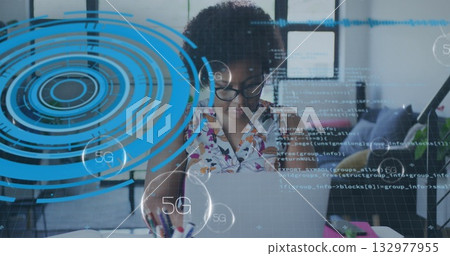 Woman in floral top focusing on laptop coding apps in modern office, with notebooks, 5G overlays 132977955