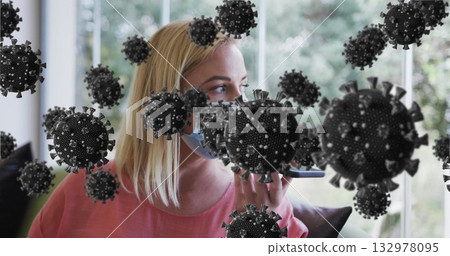 Woman wearing mask, salmon top, speaking into smartphone on sofa by window, with 3D virus particles 132978095