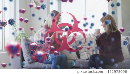 Chatting mother and daughter holding mugs in living room, floating virus graphics, biohazard symbol 132978096