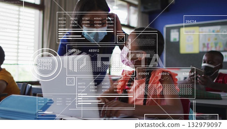 Female teacher wearing protective face mask assisting seated student in classroom, with laptops 132979097