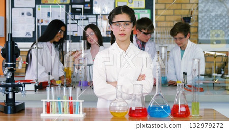 Girl wearing lab coat and goggles overseeing classmates mixing chemicals in school lab, with flasks Girl wearing lab coat and goggles overseeing classmates mixing chemicals in school lab, with flasks 132979272
