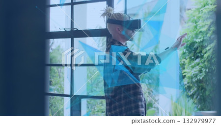 Using VR headset, male wearing dark plaid shirt gesturing in studio by window, with blue overlays 132979977