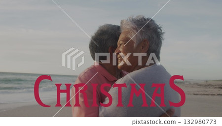 Hugging senior couple wearing pink polo on sandy beach at dusk, with red Christmas overlay 132980773