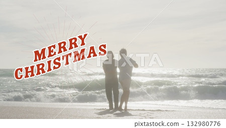 Standing senior couple watching ocean waves at sandy beach shore, with Merry Christmas overlay 132980776