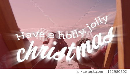 Couple in swimwear holding hands in wooden chairs on sandy shore by ocean, with Christmas overlay 132980802