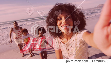 Smiling woman taking selfie on sandy beach with ocean waves at sunset, with heart graphics overlay 132980803