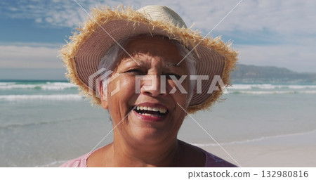 Smiling senior woman wearing straw sun hat and pink blouse standing along beach, with waves Smiling senior woman wearing straw sun hat and pink blouse standing along beach, with waves 132980816