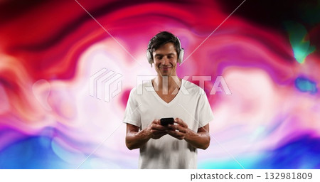 Standing man wearing white v-neck tee and headphones against abstract backdrop, holding smartphone 132981809