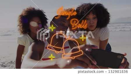 Family taking selfie on sandy beach at water's edge, with smartphone, neon Jingle Bells overlay 132981836