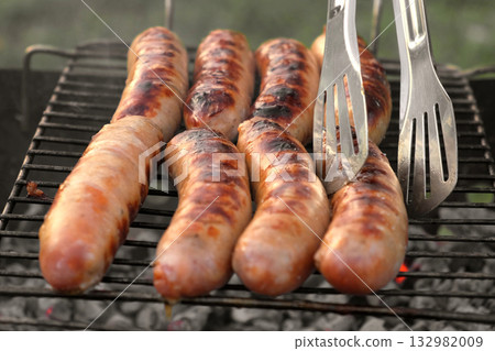 Grilled sausages. Cook man preparing grilling food bbq. Barbecue with smoke, flame outdoors. Tasty juicy german bratwurst. Charcoal kettle grill outside in backyard. Family summer vacation. Close-up 132982009
