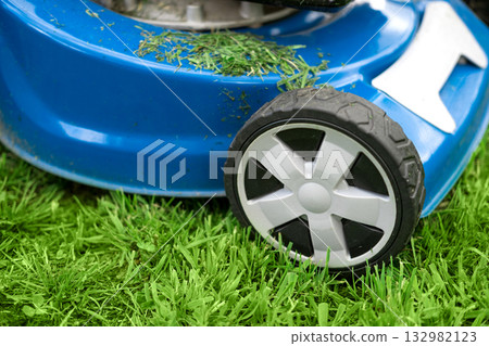 Lawn mower cutting grass. Small grass cuttings fly out of lawnmower. Grass clippings get spewed out of a mower pushed around by landscaper. CloseUp. Gardener working with mower machine. Mowing lawns Lawn mower cutting grass. Small grass cuttings fly out of lawnmower. Grass clippings get spewed out of a mower pushed around by landscaper. CloseUp. Gardener working with mower machine. Mowing lawns 132982123