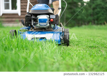 Lawn mower cutting grass. Small grass cuttings fly out of lawnmower. Grass clippings get spewed out of a mower pushed around by landscaper. CloseUp. Gardener working with mower machine. Mowing lawns Lawn mower cutting grass. Small grass cuttings fly out of lawnmower. Grass clippings get spewed out of a mower pushed around by landscaper. CloseUp. Gardener working with mower machine. Mowing lawns 132982125