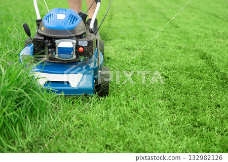 Lawn mower cutting grass. Small grass cuttings fly out of lawnmower. Grass clippings get spewed out of a mower pushed around by landscaper. CloseUp. Gardener working with mower machine. Mowing lawns Lawn mower cutting grass. Small grass cuttings fly out of lawnmower. Grass clippings get spewed out of a mower pushed around by landscaper. CloseUp. Gardener working with mower machine. Mowing lawns 132982126
